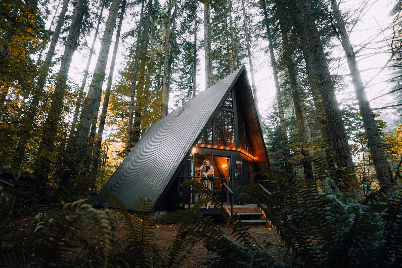 A-frame cabin nestled in a dense forest with tall, towering trees. Warm lights illuminate the cabin's entrance, creating a cozy, inviting atmosphere.