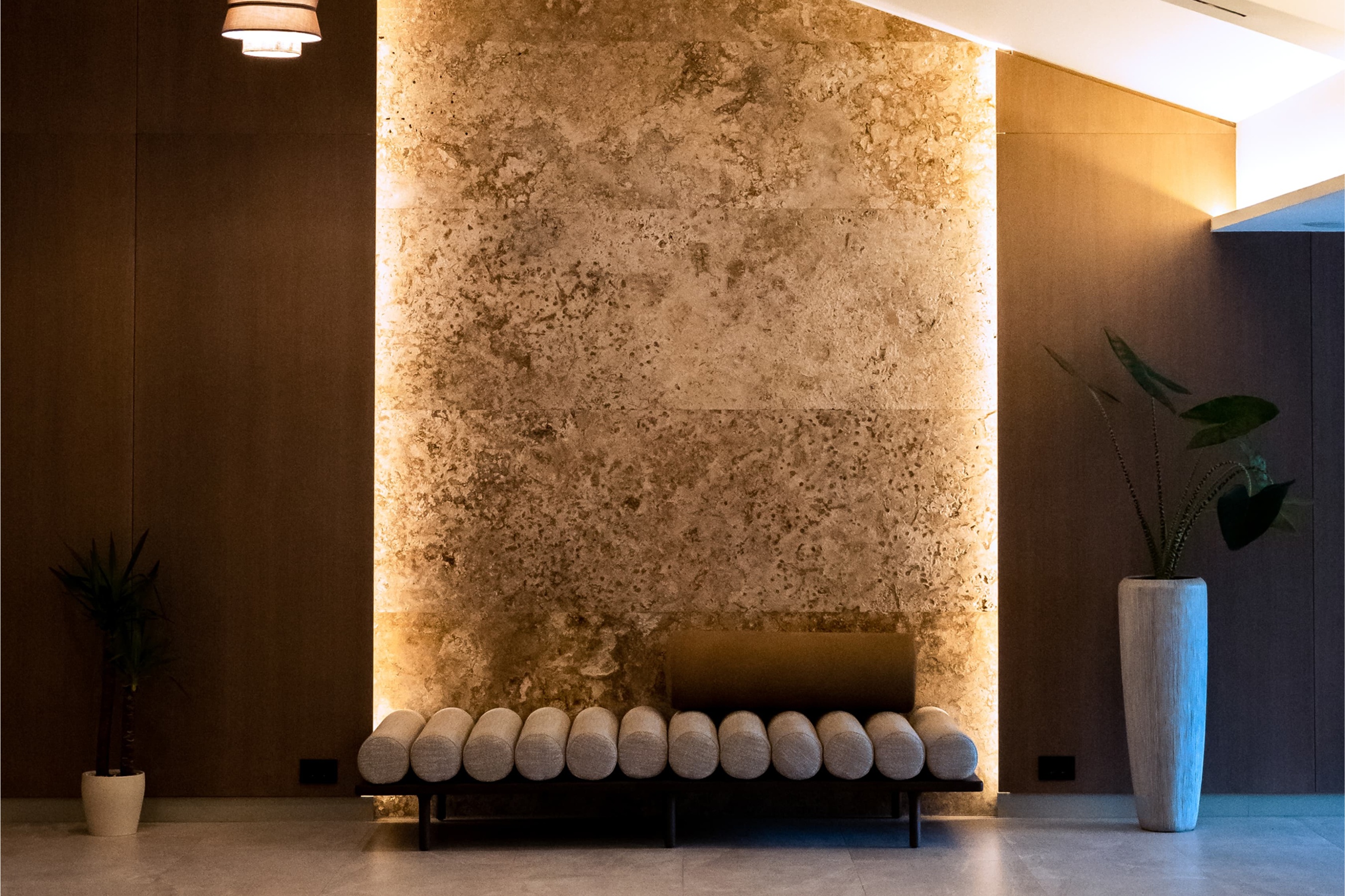 Modern interior with a minimalist bench in front of a textured, backlit stone wall. Warm lighting adds a calming ambiance in known wellness & spa destination in Serbia, Vrujci spa known for natural spa experience. Plants in the corners add freshness.
