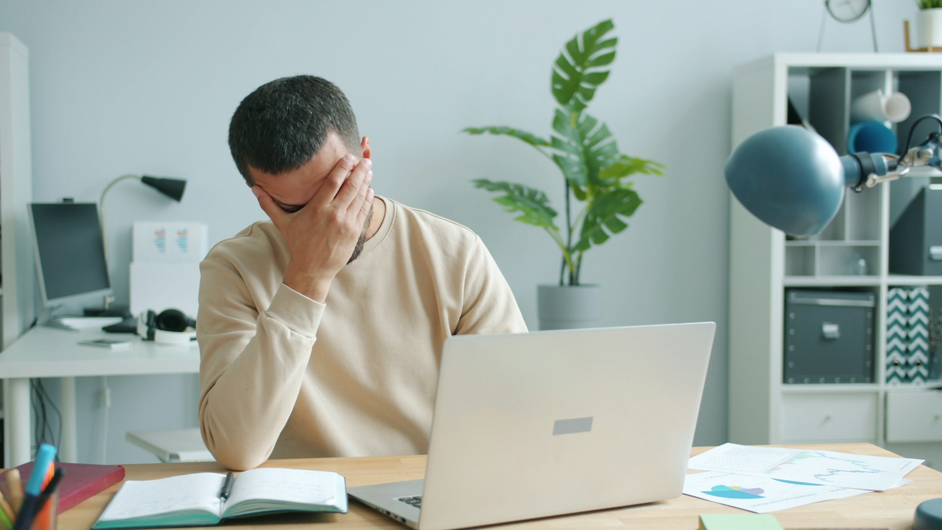 Man in a beige sweater sits at a desk with a laptop reflecting burnout situation. He covers his face with his hand, suggesting stress or frustration. A plant and papers are visible.