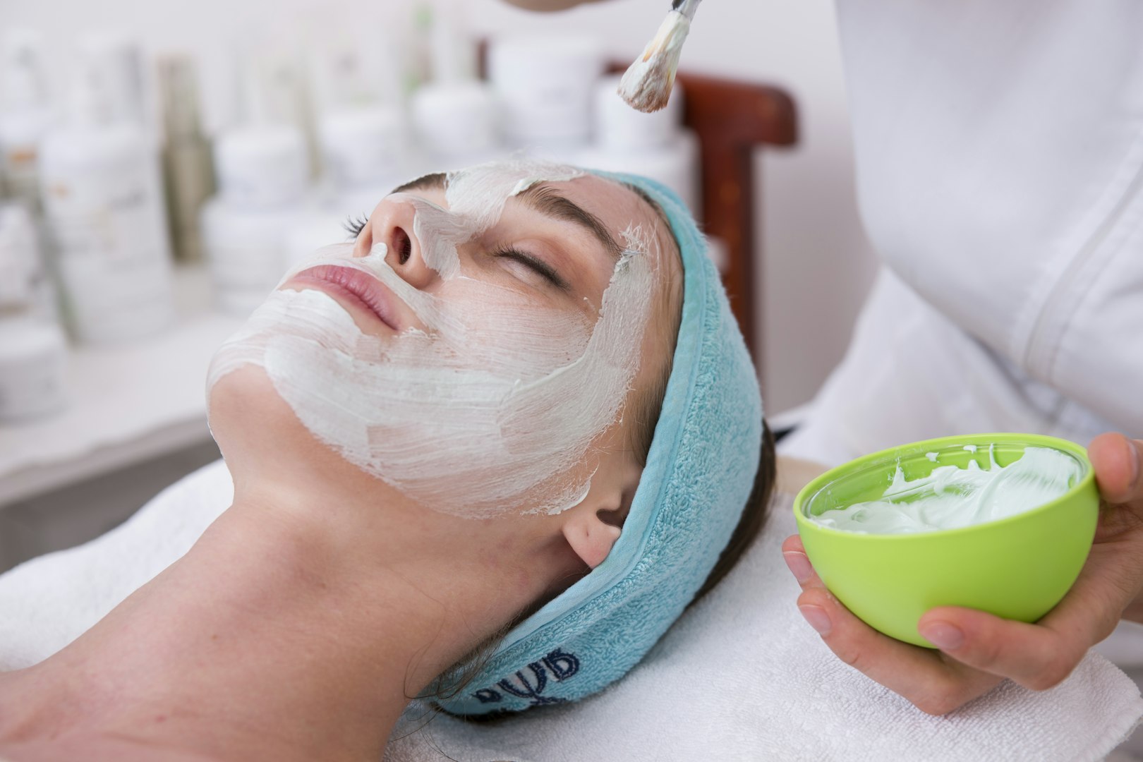 A person receives a relaxing facial treatment, eyes closed, with a white mask applied by a specialist holding a bowl. The atmosphere is calm and soothing.