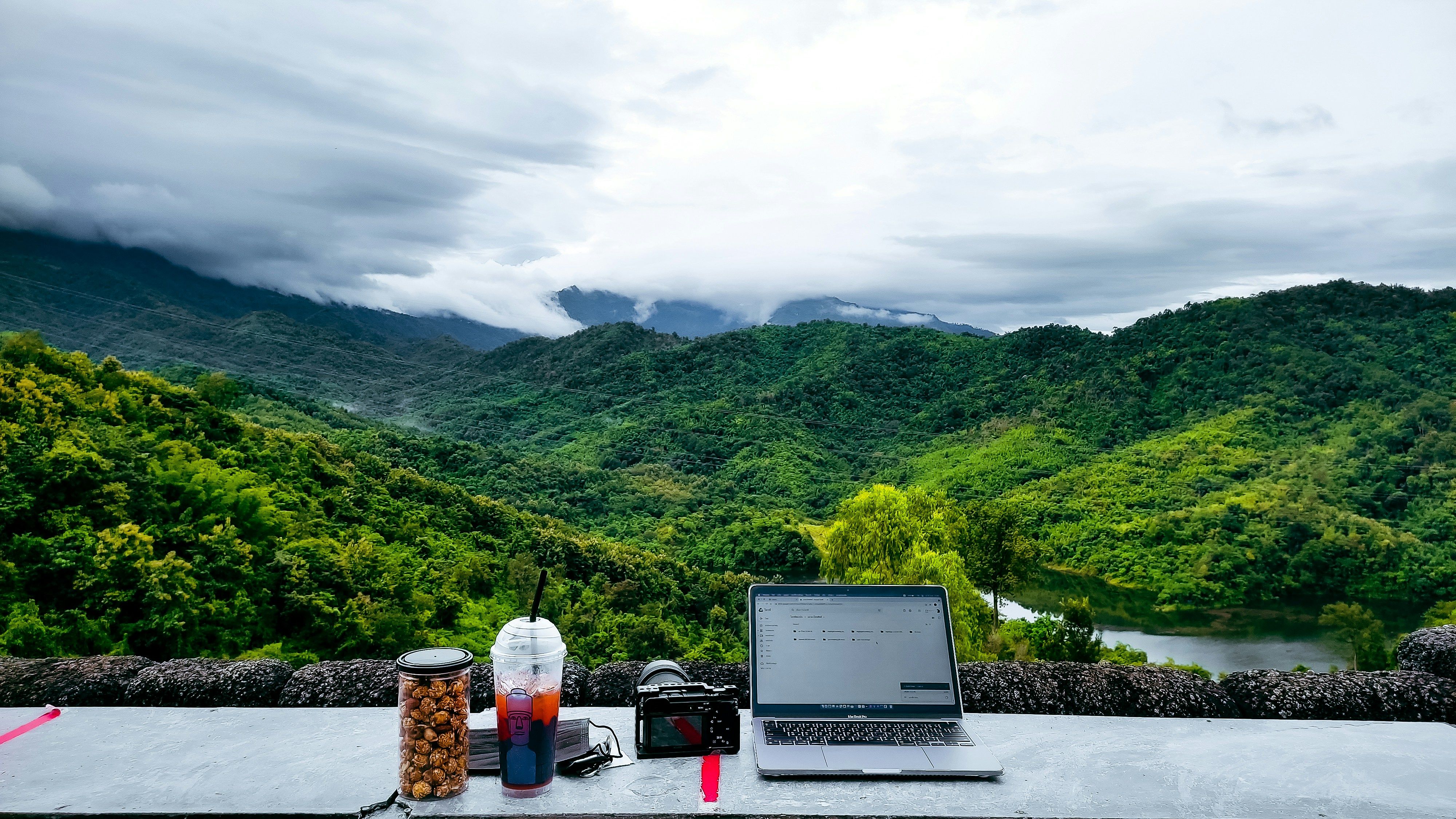 Laptop on a stone table with snacks and camera, overlooking lush green hills and cloudy sky. A serene remote work setup, blending nature and technology into digital detox philosophy.