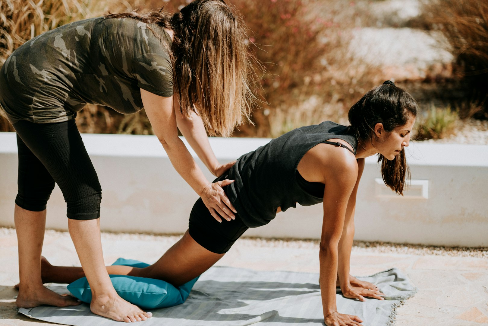 A woman assists another in a yoga pose outdoors. The assisting woman, wearing a camouflage shirt, supports the other’s hips as she stretches on a mat. Calm and focused ambiance.