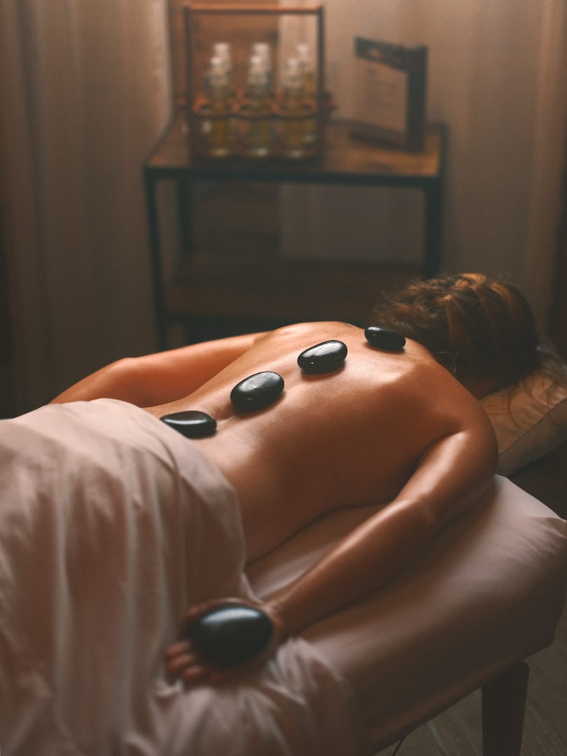 A woman is lying face down on a massage table under a white sheet, with smooth, dark massage stones placed along her back. The room is softly lit, giving a calming and serene atmosphere. In the background, a table holds various bottles, adding to the spa-like setting, perfect for massage and spa centre recommendations.