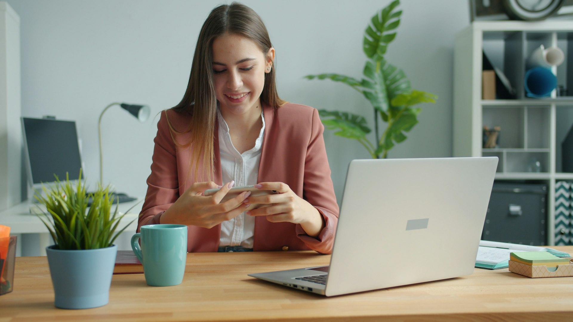 A woman in a pink blazer smiles while looking at her phone, seated at a wooden desk with a laptop, coffee mug, and a plant in a bright office.